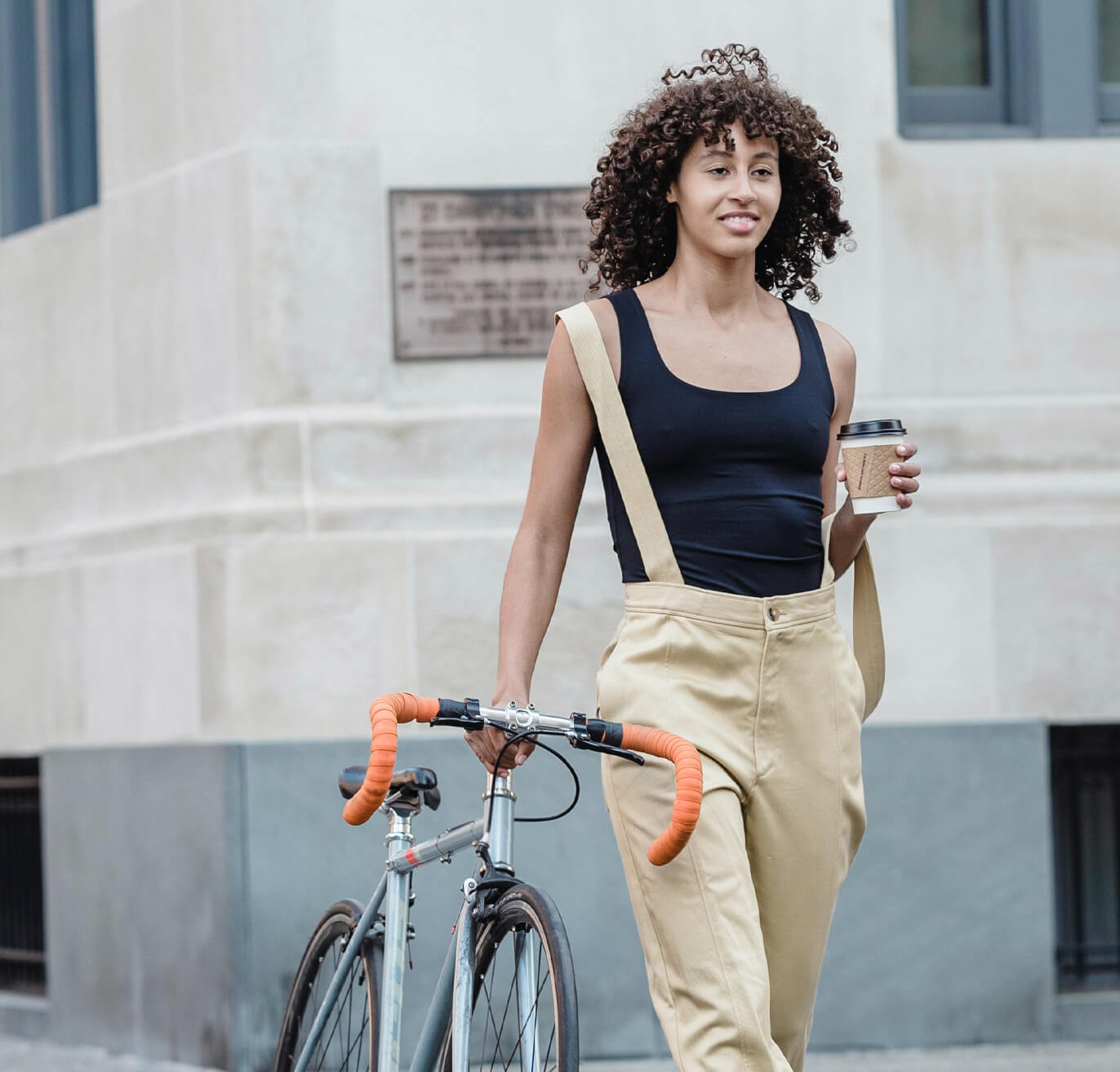 A woman walking holding her bike and a coffee.